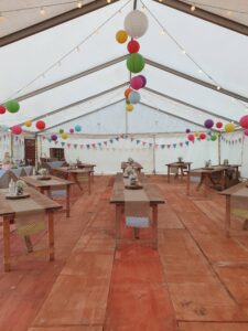 Inside of marquee decorated with blue and pink bunting and bright colourful paper lanterns. In the distance on the left is the card collecting box and the cake table and a donut wall. Each rustic rectangular trestle table has a hessian table runner, a log slice, a jar of sweets, a bottle with fairy lights in and a painted can with gypsophila