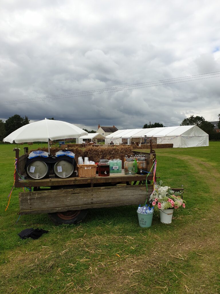 The white marquee is in the background and in the foreground is a rustic wooden trailer with two kegs of beer/lager and jugs of gn