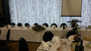 long top table with chairs  in front of a bright white light up curtain and projector screen